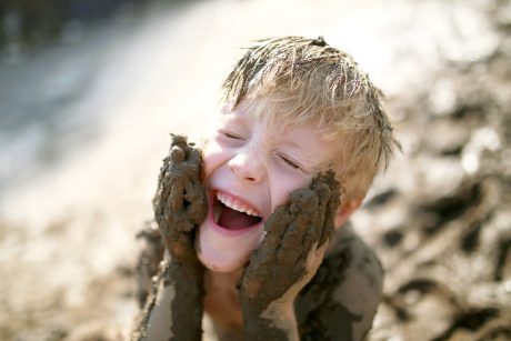 A cute little boy child is laughing as he plays outside in the mud and rubs dirt on his face with his hands.
